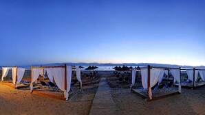 A beach with wooden cabanas, white drapes, and a wooden walkway leading to the water.