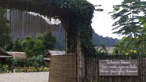 Una rústica puerta de madera con un letrero que indica Phoxay Ngam Coffee & Restaurant, rodeada de exuberante vegetación y un telón de fondo montañoso.