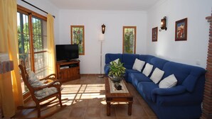A living room with a blue sofa, a wooden rocking chair, a television on a wooden stand, a floor lamp, and a potted plant on a wooden table. - Belvilla by OYO Francisco (Frigiliana)