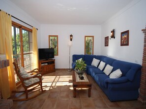 Un salon avec un canapé bleu, un fauteuil à bascule en bois, un téléviseur posé sur un meuble en bois, une lampe sur pied et une plante en pot sur une table en bois.