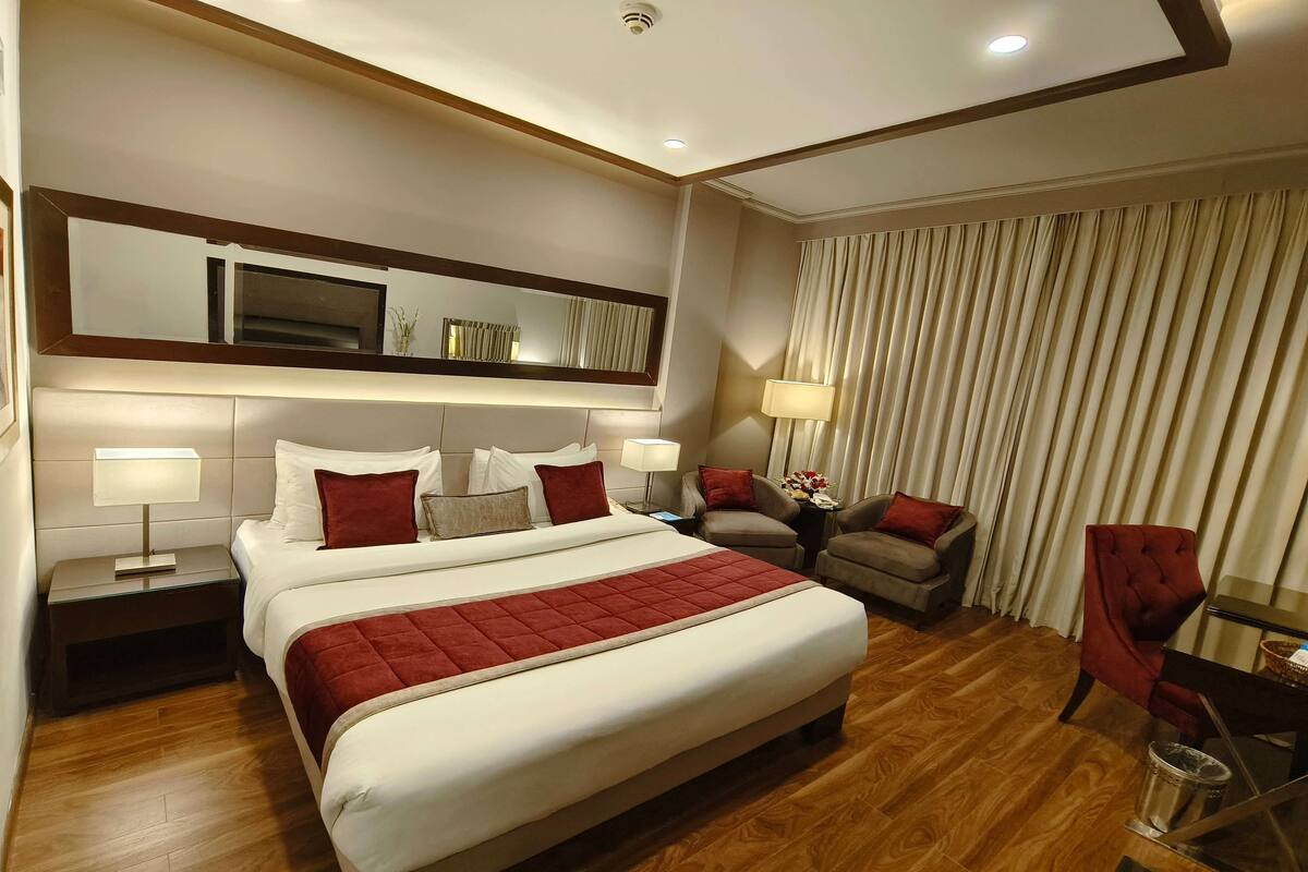 Une chambre d’hôtel avec un grand lit, une table de chevet avec une lampe, une chaise, une petite table et un rideau.
