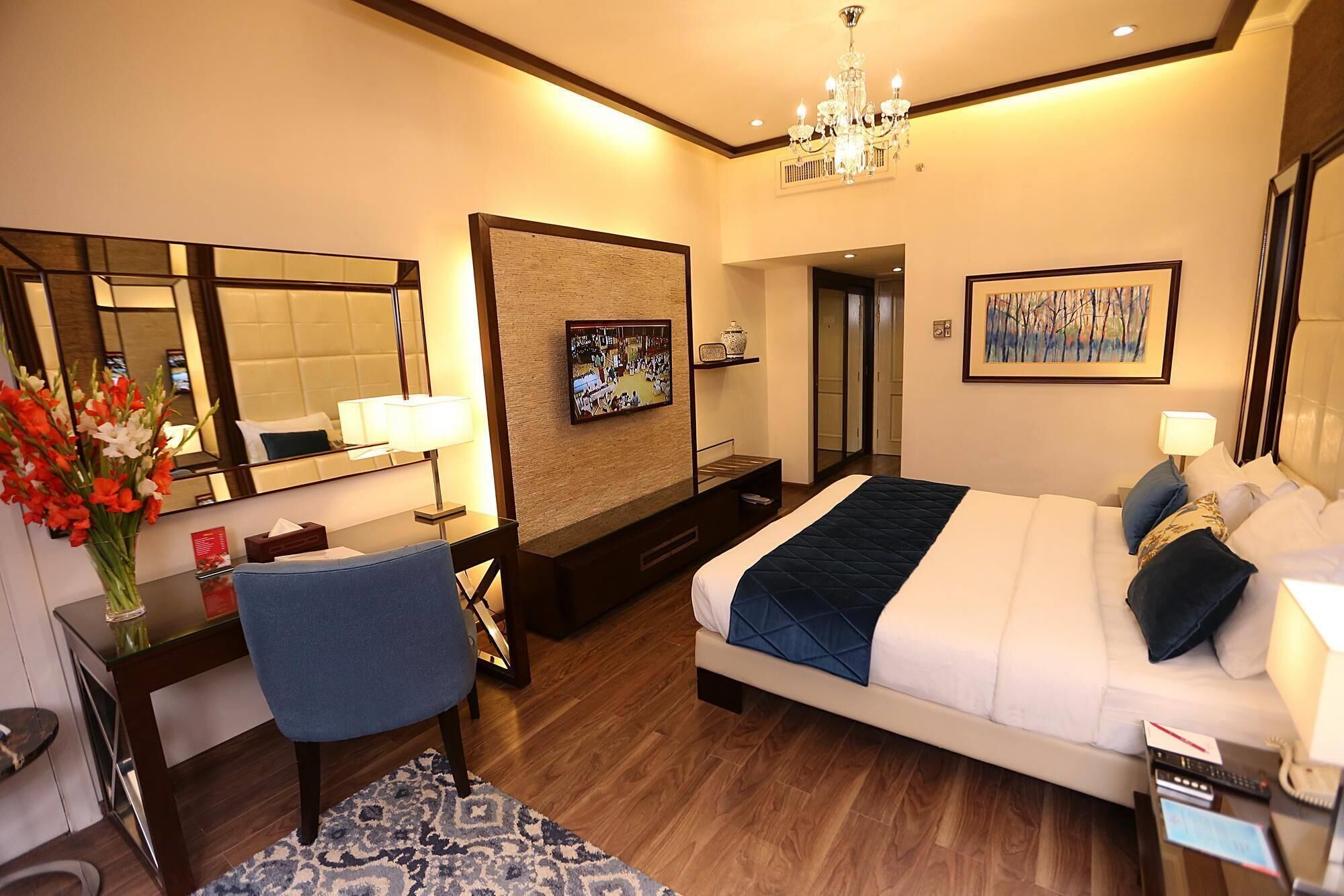 A hotel room with a large bed, a desk with a chair, a TV, a mirror, and a chandelier.