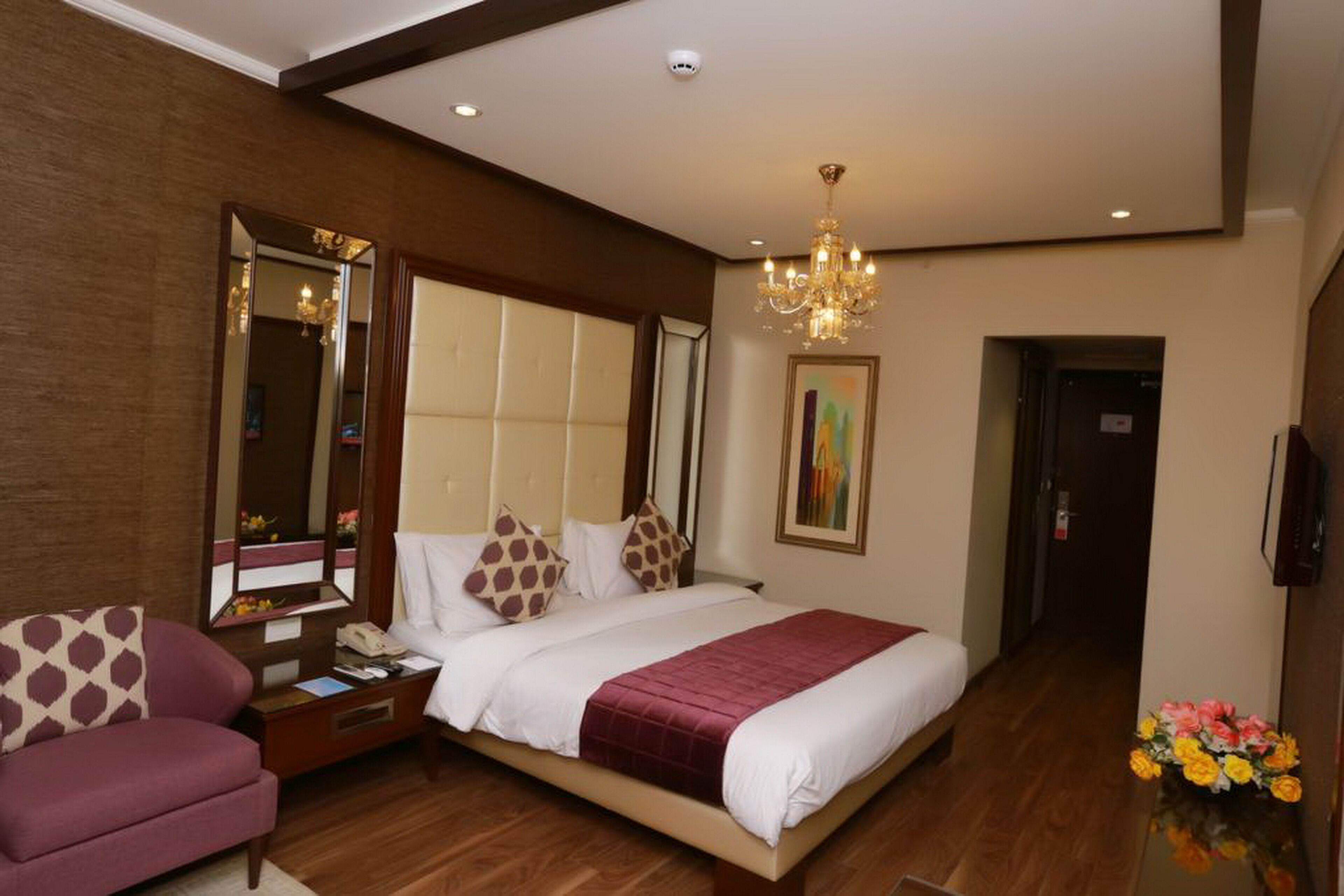 A hotel room with a large bed, a sofa, a nightstand, a mirror, a chandelier, and a painting on the wall.