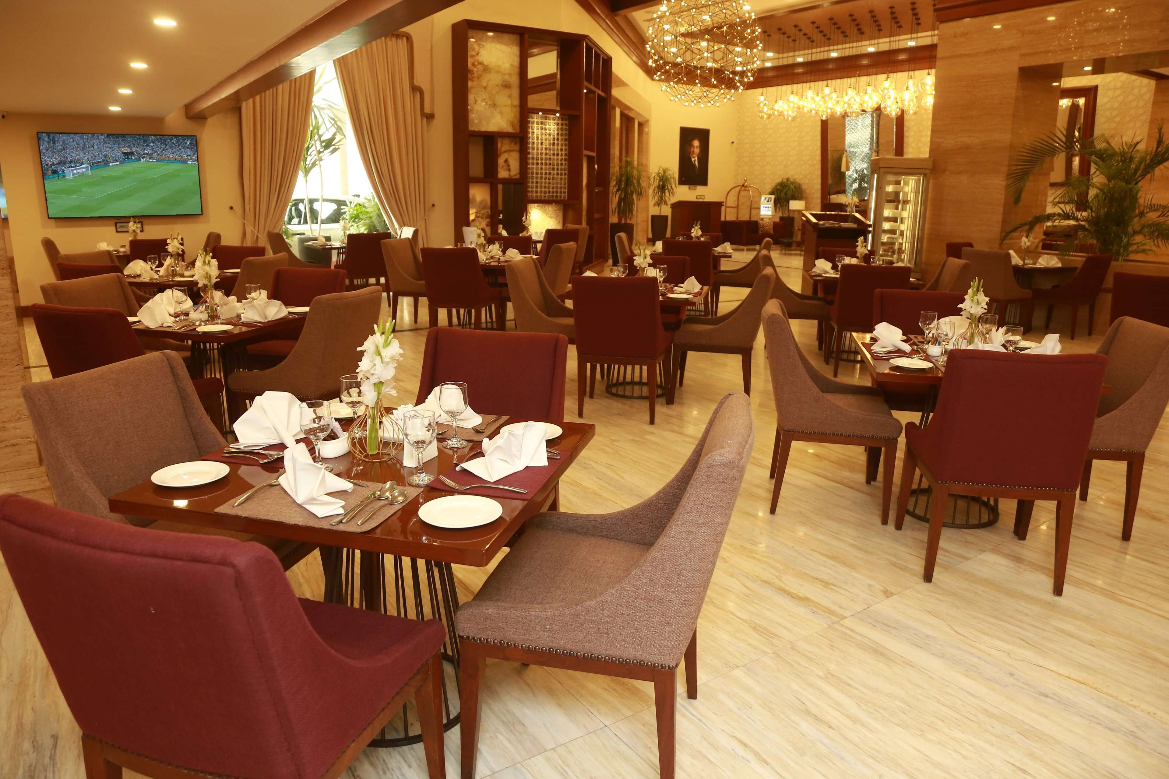 Une salle à manger d’hôtel avec de nombreuses tables dressées, des chaises et un grand écran diffusant un événement sportif.