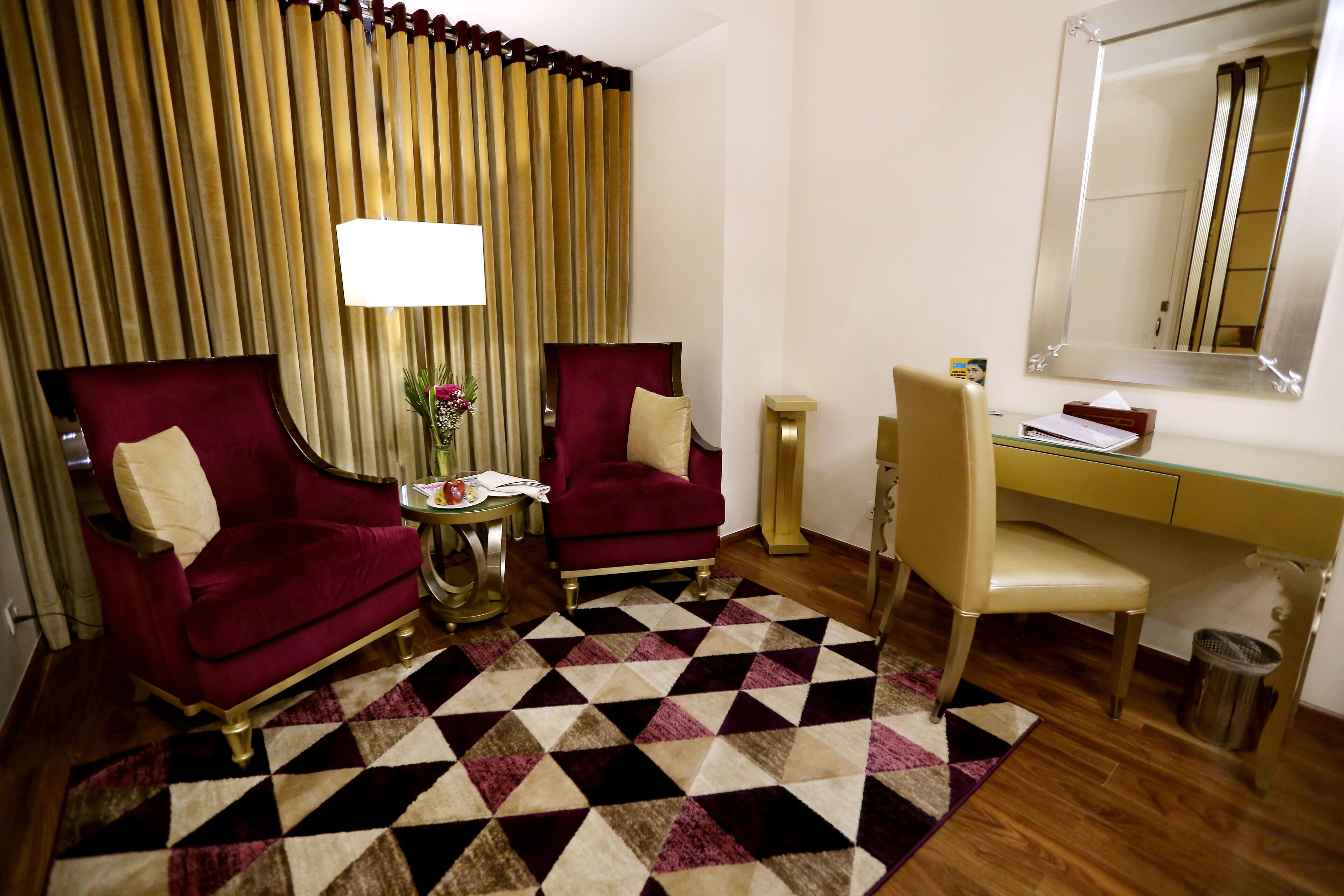 Ein Hotelzimmer mit zwei roten Sesseln, einem beigen Stuhl, einem Schreibtisch, einem Spiegel und einem gemusterten Teppich.
