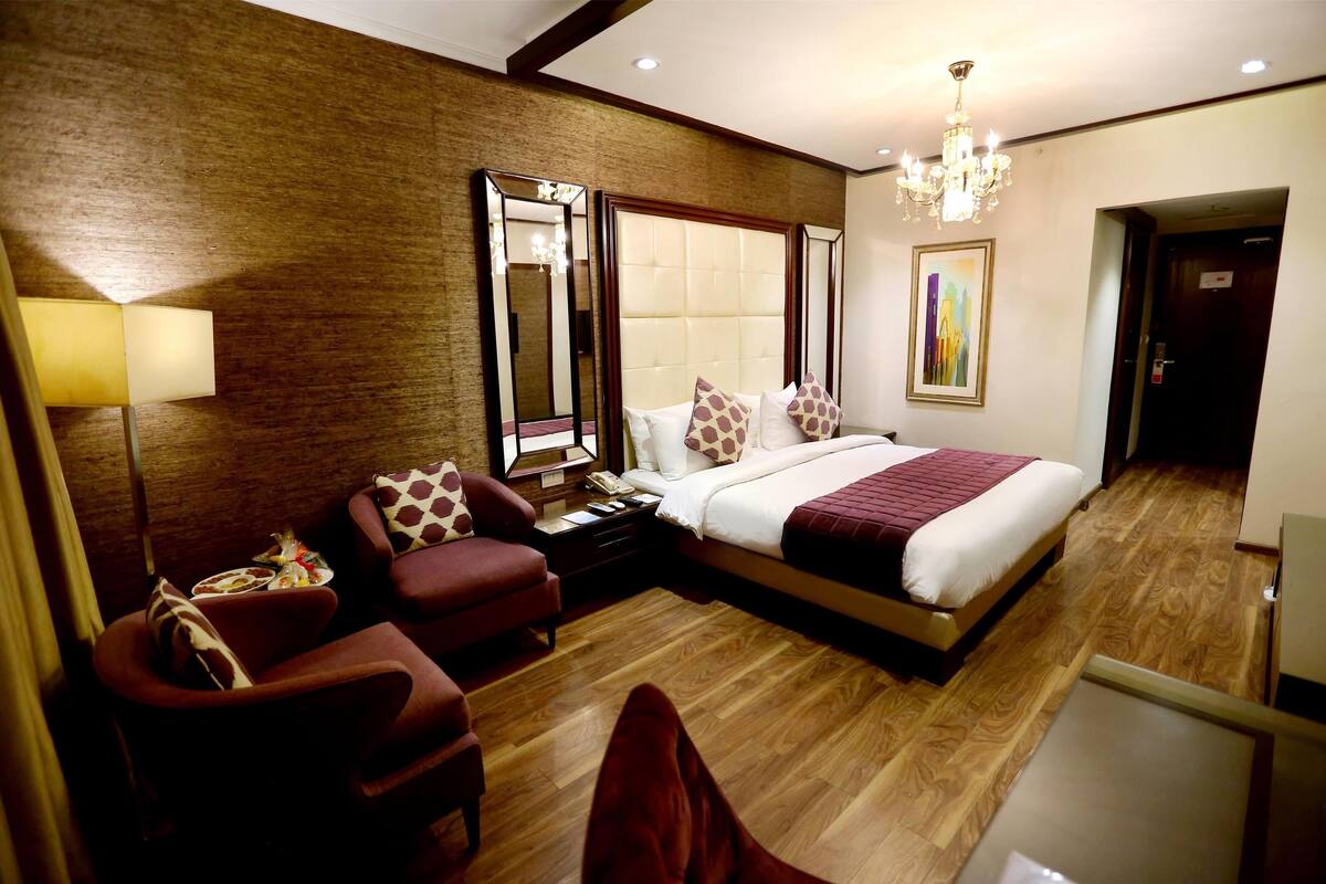 Une chambre d’hôtel avec un grand lit, un coin salon avec un fauteuil, une table de chevet, une lampe et un tableau au mur.