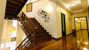 A wooden staircase with a chandelier, a wall with decorative plates, and a hallway with a polished floor. - Srisomboon Hostel (Trang)