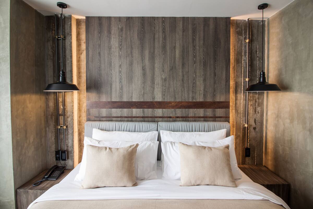 A modern hotel room with a wooden headboard, a large bed with white linens and beige pillows, and two bedside tables with black lamps.