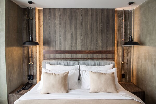 A modern hotel room with a wooden headboard, a large bed with white linens and beige pillows, and two bedside tables with black lamps.