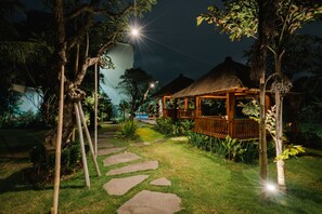 Un jardín nocturno con un pabellón de techo de paja, senderos de piedra y vegetación tropical.