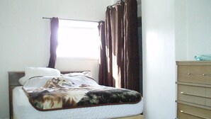 A bed with a patterned blanket, a wooden dresser, and a window with curtains. - Canora Cozy Cottage (Canora)