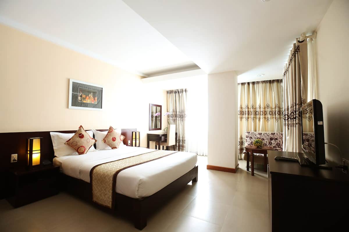 A hotel room with a large bed, a desk, a chair, a TV, and a small table with a flower vase.