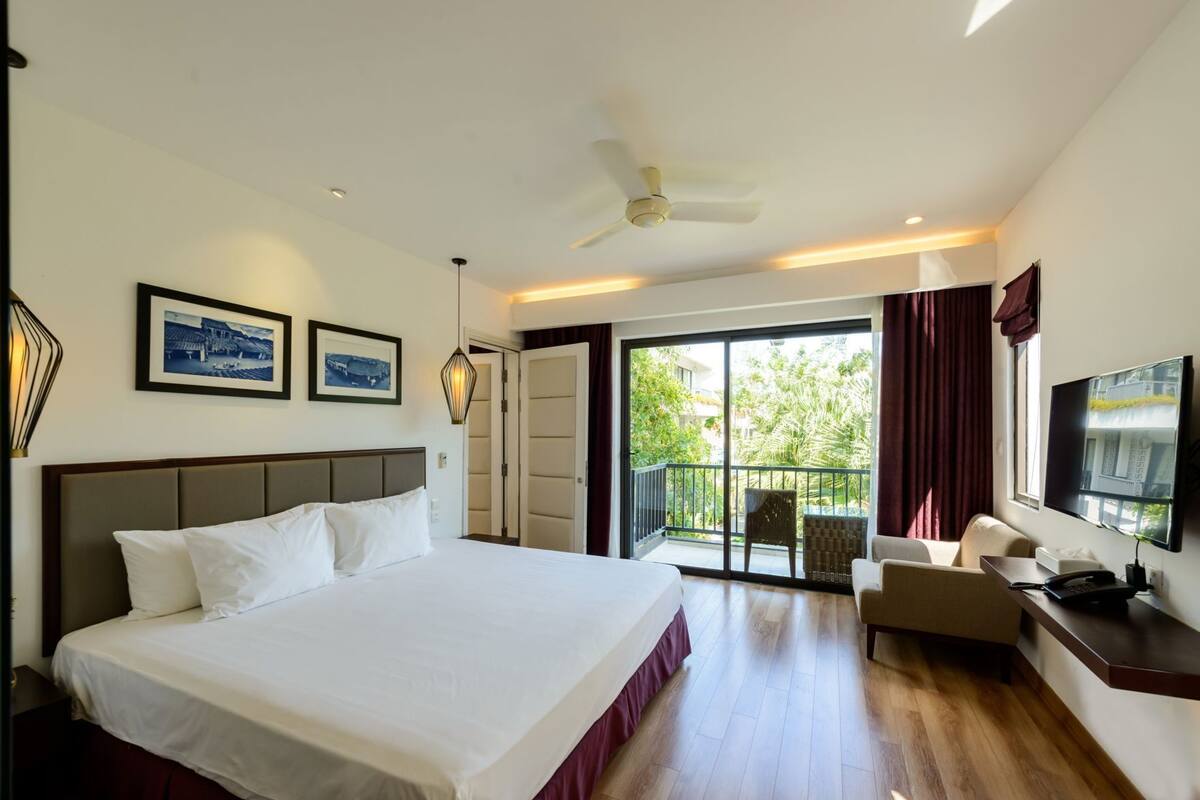 A hotel room with a large bed, a desk, a chair, a TV, and a balcony with a view of greenery.