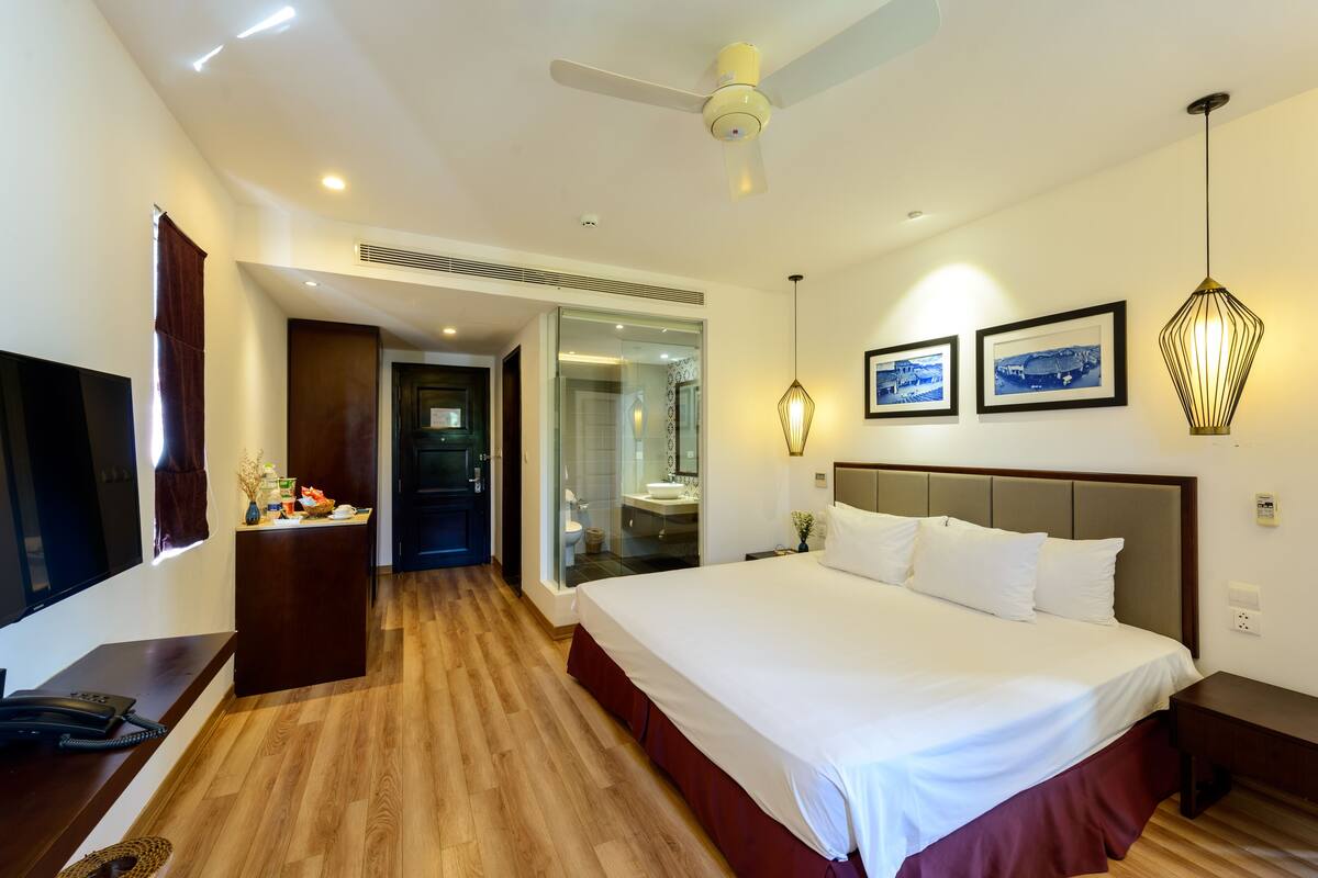 A hotel room with a large bed, a desk with a telephone, a TV, and a ceiling fan.