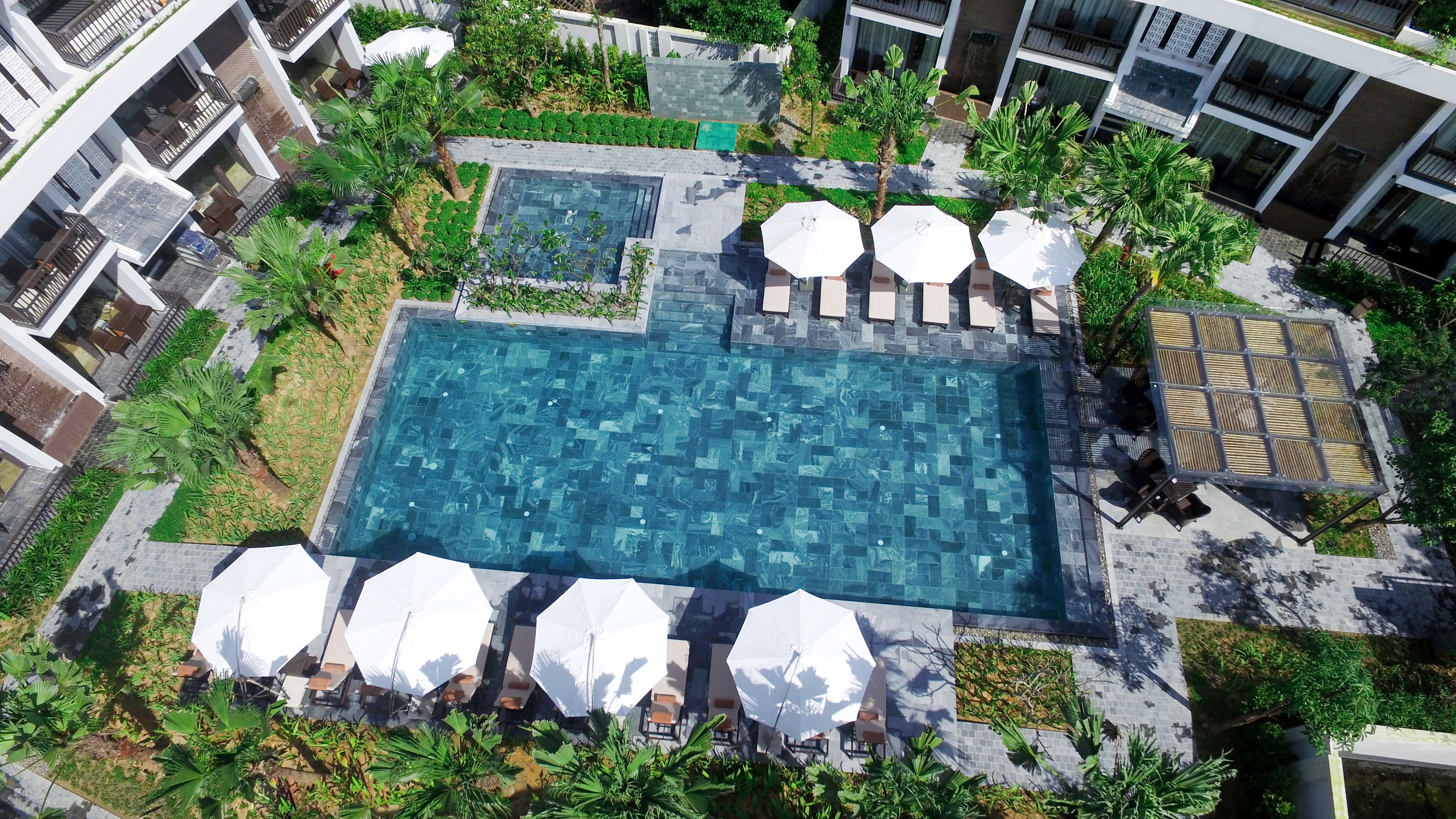 An aerial view of a modern hotel with a central swimming pool, surrounded by lush greenery and outdoor seating areas.