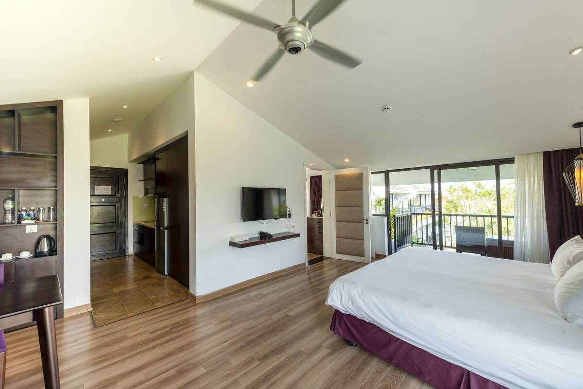 A modern hotel room with a bed, a TV, a balcony with a view, and a kitchen area.