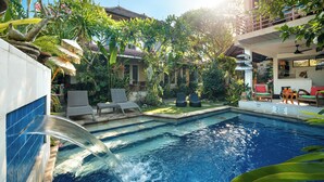 A pool with a waterfall feature, surrounded by lush greenery and a two-story building with outdoor seating. - Angel House Ubud Eco Villa (Mas)