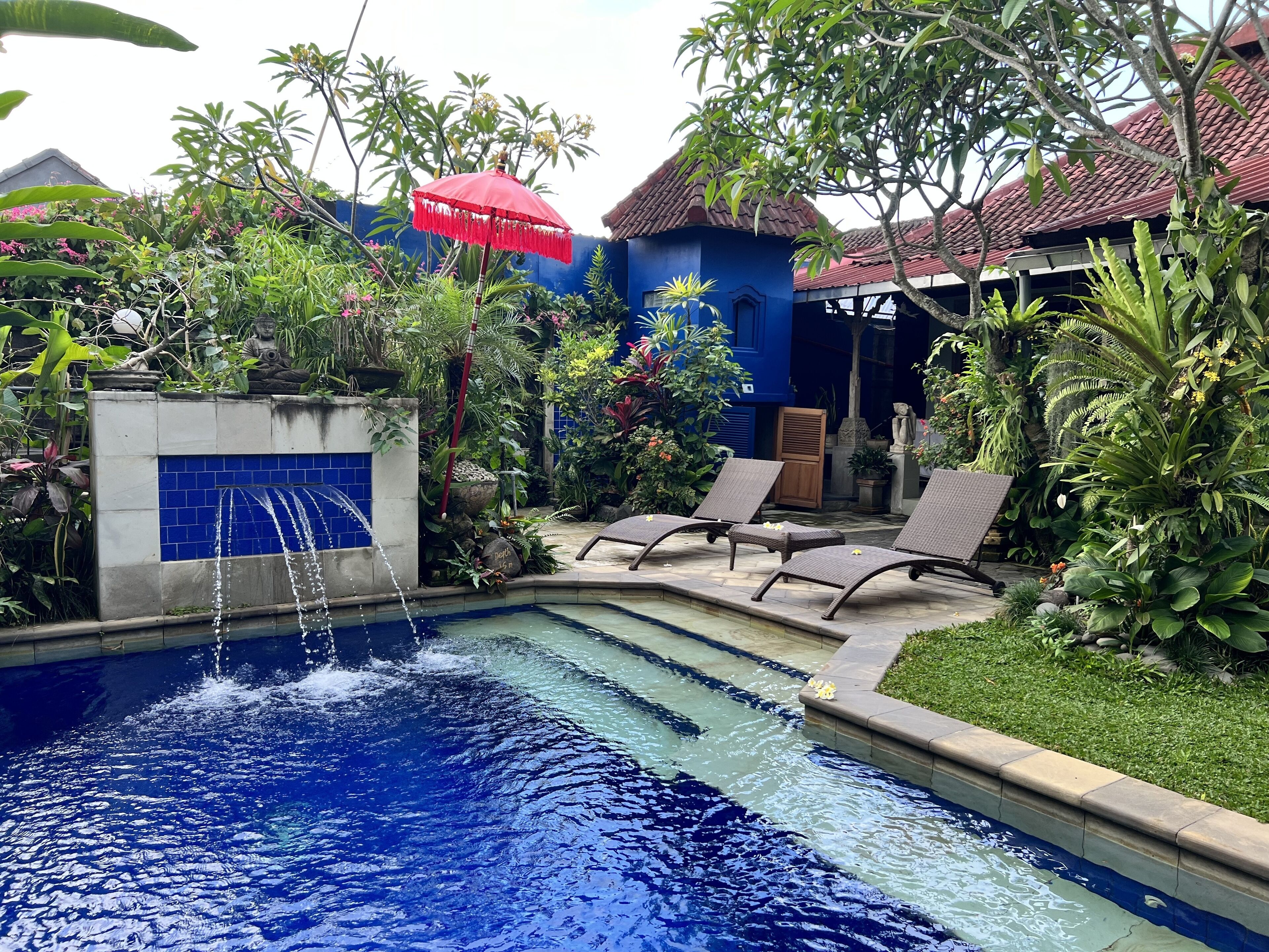 A swimming pool with a water fountain, surrounded by lush greenery and a blue building.