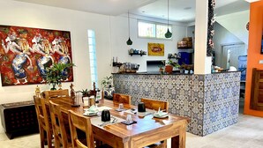 A dining area with a wooden table set for six, surrounded by chairs. A kitchen is visible in the background with a tiled backsplash and various kitchen items on the counter. - Angel House Ubud Eco Villa (Mas)