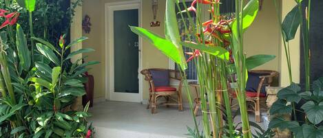 A tropical garden entrance with a door, wicker chairs, and various plants.