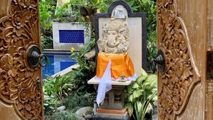 A wooden door with intricate carvings, leading to a courtyard with a statue, plants, and a tiled wall. - Angel House Ubud Eco Villa (Mas)