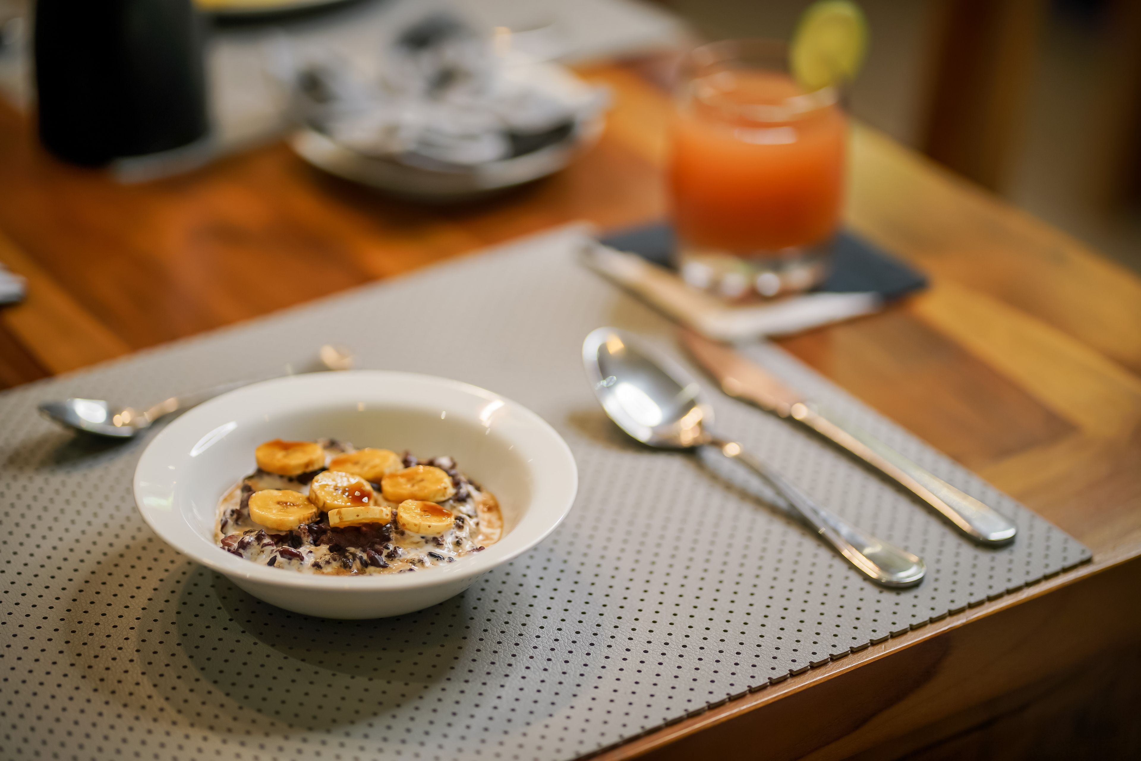 Une table en bois dressée pour le petit-déjeuner, avec un bol de céréales garni de bananes et un verre de jus d’orange.