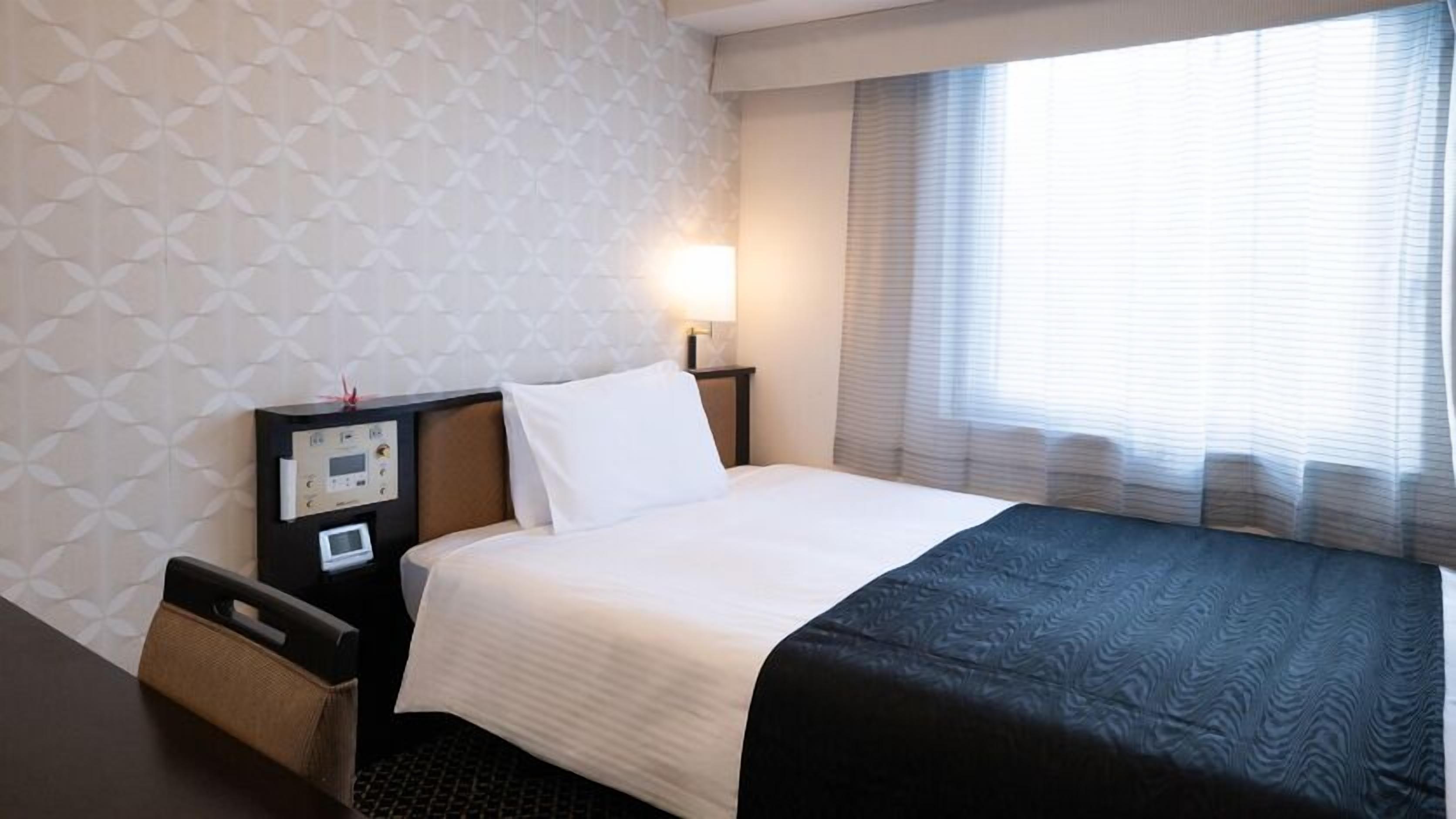 Quarto de hotel com cama, mesa de cabeceira, luminária, relógio de parede e janela com persianas.