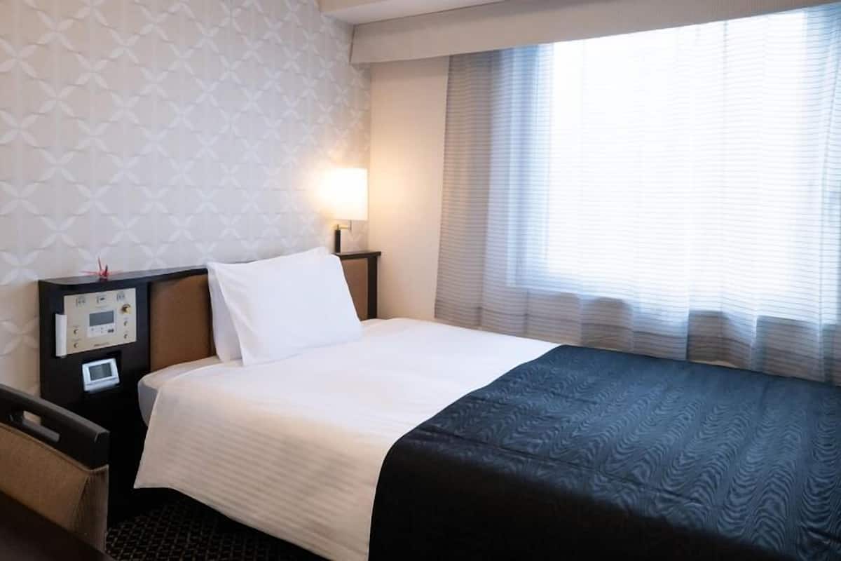 Habitación de hotel con cama, mesita de noche, lámpara, reloj de pared y ventana con persianas.