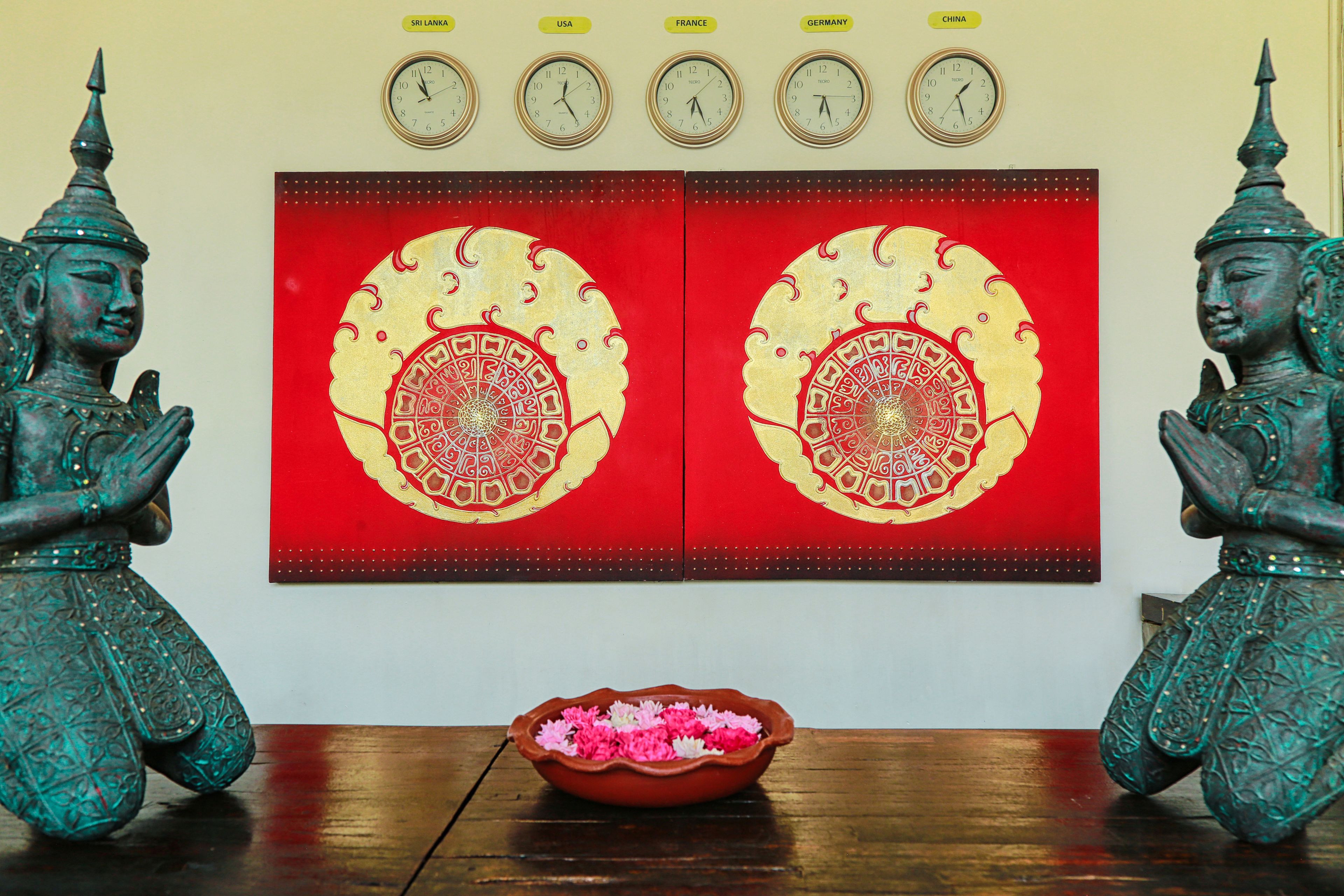 A room with two Buddha statues, a red decorative panel with intricate designs, a clock above the panel, and a bowl of flowers on the table.