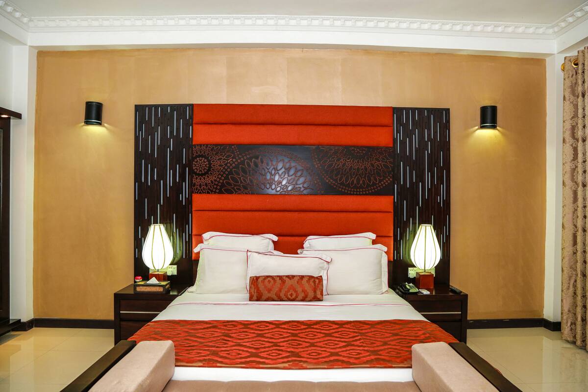 A hotel room with a large bed, two bedside tables with lamps, a decorative headboard, and a patterned throw blanket.