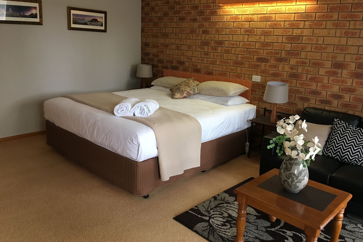 Habitación de hotel con pared de ladrillo, una cama con ropa blanca, dos cuadros en la pared, una mesita de noche con una lámpara, un sofá de cuero negro y un jarrón con flores.