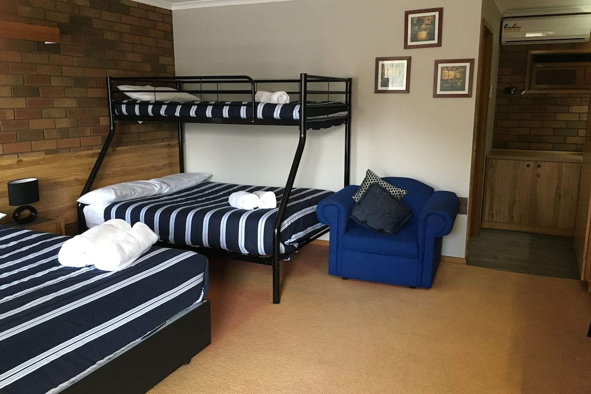 Una habitación con dos literas, un sillón azul, una mesita con una lámpara y una pared de ladrillo.