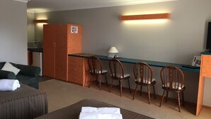 Une chambre d’hôtel compacte dotée d’une kitchenette, d’un coin repas avec quatre chaises en bois, d’un canapé et d’un lit soigneusement fait, avec une serviette blanche pliée.