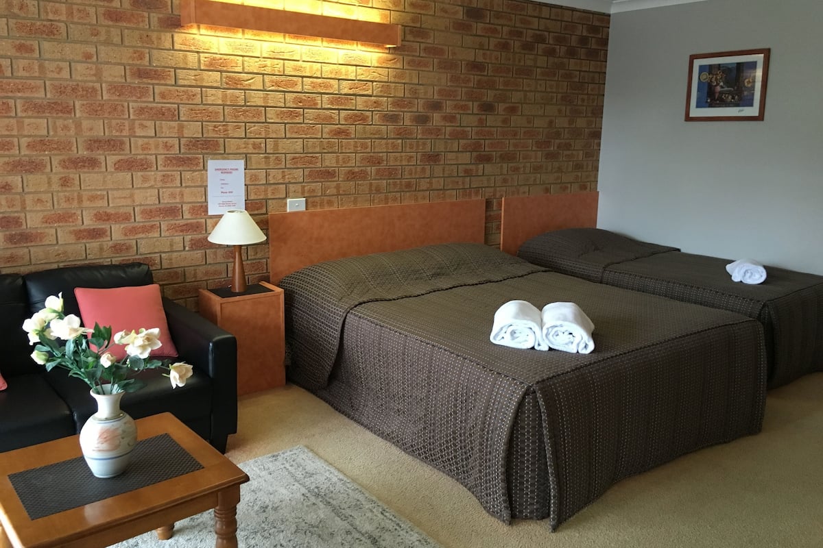 Habitación de hotel con pared de ladrillo, dos camas, un sofá, una mesita de noche, una lámpara, un jarrón con flores y un cuadro en la pared.