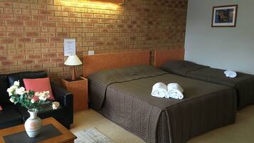 A hotel room with a brick wall, two beds, a sofa, a nightstand, a lamp, a vase with flowers, and a framed picture on the wall.