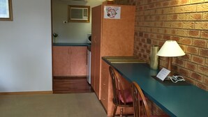 Une petite cuisine équipée d’un réfrigérateur, d’un four à micro-ondes et d’un évier. On y trouve deux chaises en bois avec des coussins roses et une table avec une lampe. La pièce est dotée d’un mur en briques et d’un sol recouvert de moquette.