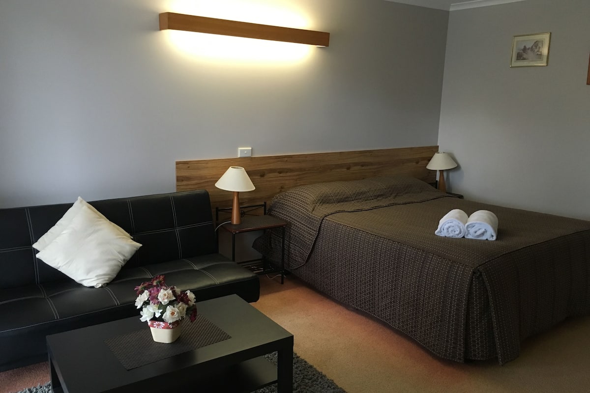 Habitación de hotel con cama, sofá, una mesita con un florero y una mesita de noche con lámpara.