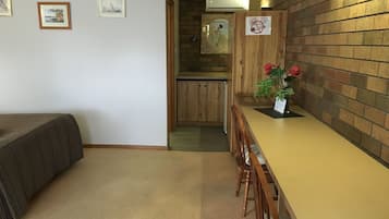 A hotel room with a bed, a wooden table with chairs, a brick wall, and a kitchenette in the background.