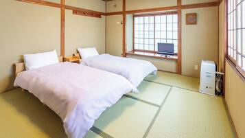 A traditional Japanese room with tatami flooring, two beds with white bedding, a desk with a computer, and a window with a view.
