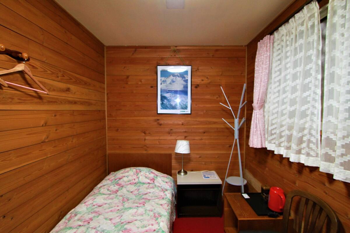 Une petite pièce chaleureuse aux murs en bois, avec un lit recouvert d’une literie à motifs floraux, une table de chevet avec une lampe, une chaise et une fenêtre ornée de rideaux légers.