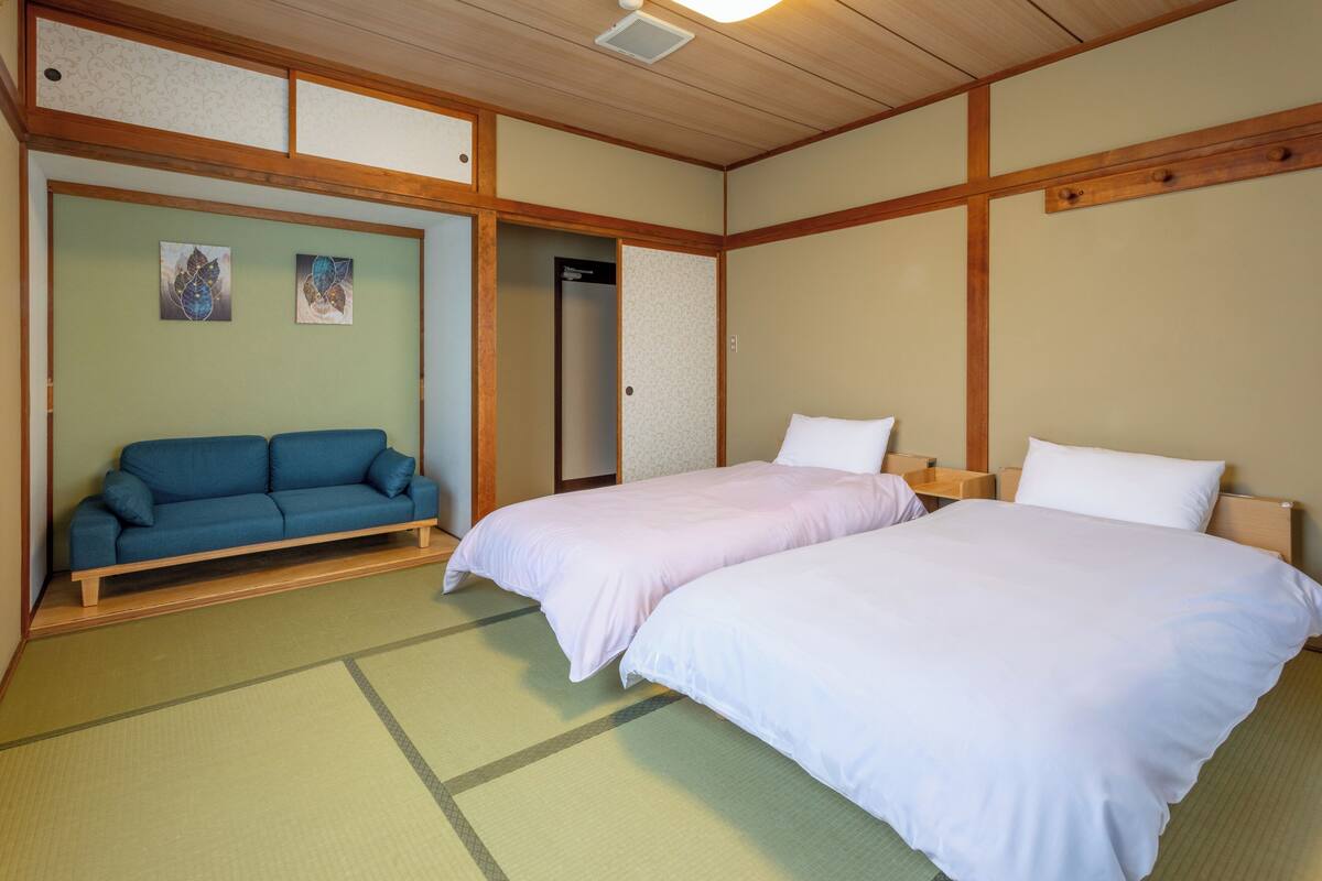 Une pièce de style japonais traditionnel, avec un sol recouvert de tatamis, un canapé bleu, deux lits avec du linge de lit blanc et des œuvres d’art accrochées aux murs.