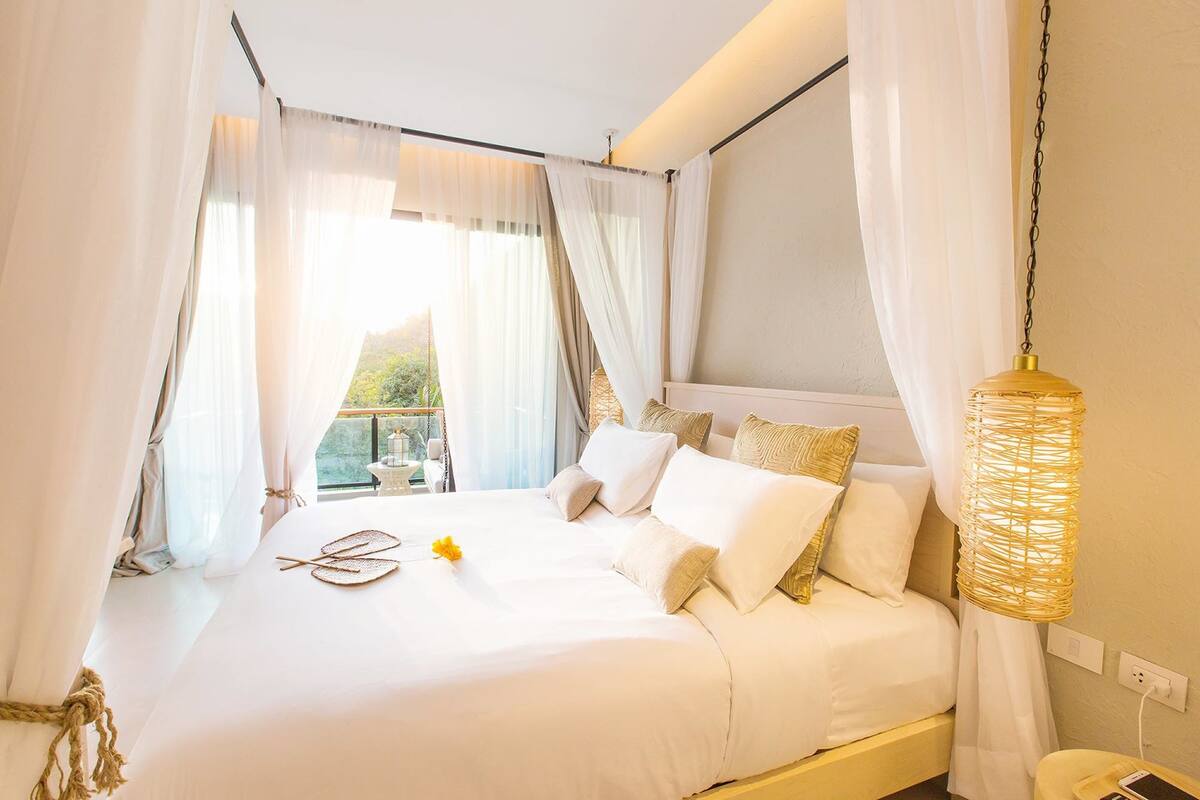 Una cama bien hecha con ropa blanca y una flor amarilla, un colgante de luz tejido y cortinas translúcidas.