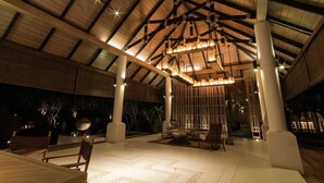 An indoor covered area with a high ceiling, wooden beams, and a central light fixture. There are seating arrangements and a decorative wall with vertical elements. - The Series Resort Khaoyai (Pak Chong)