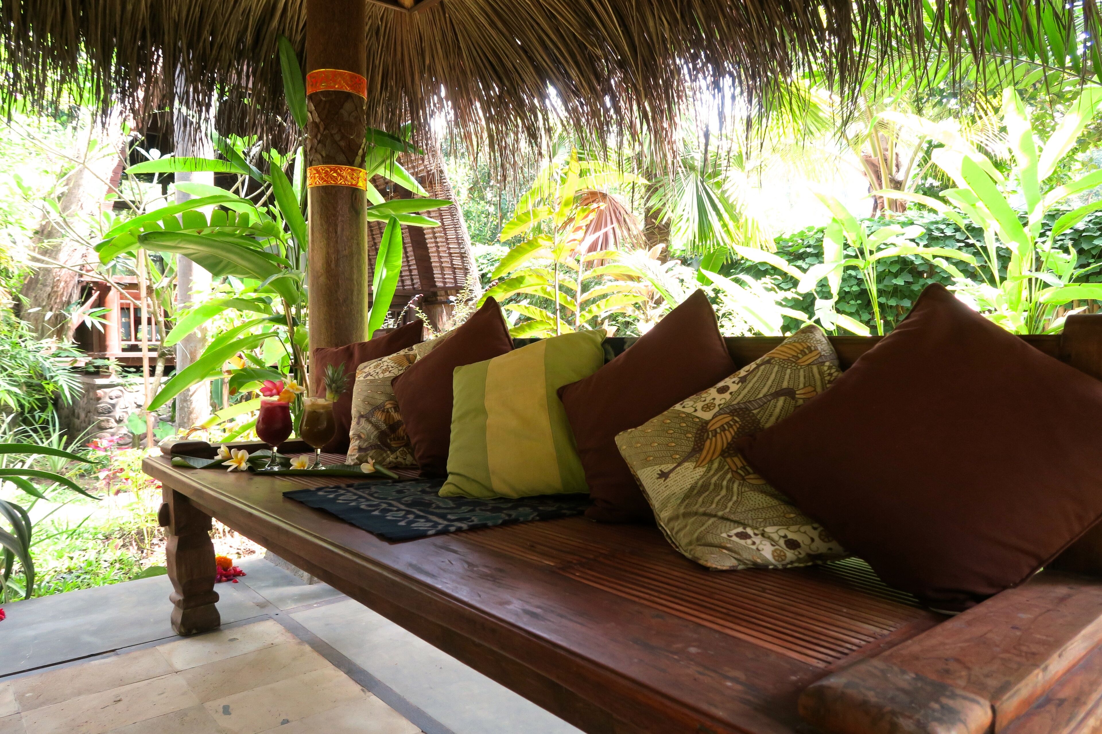 A wooden bench with cushions and a woven mat, surrounded by lush tropical plants.