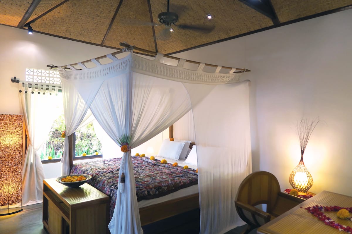 A room with a bed under a canopy, a wooden nightstand, a chair, and a table with a lamp.