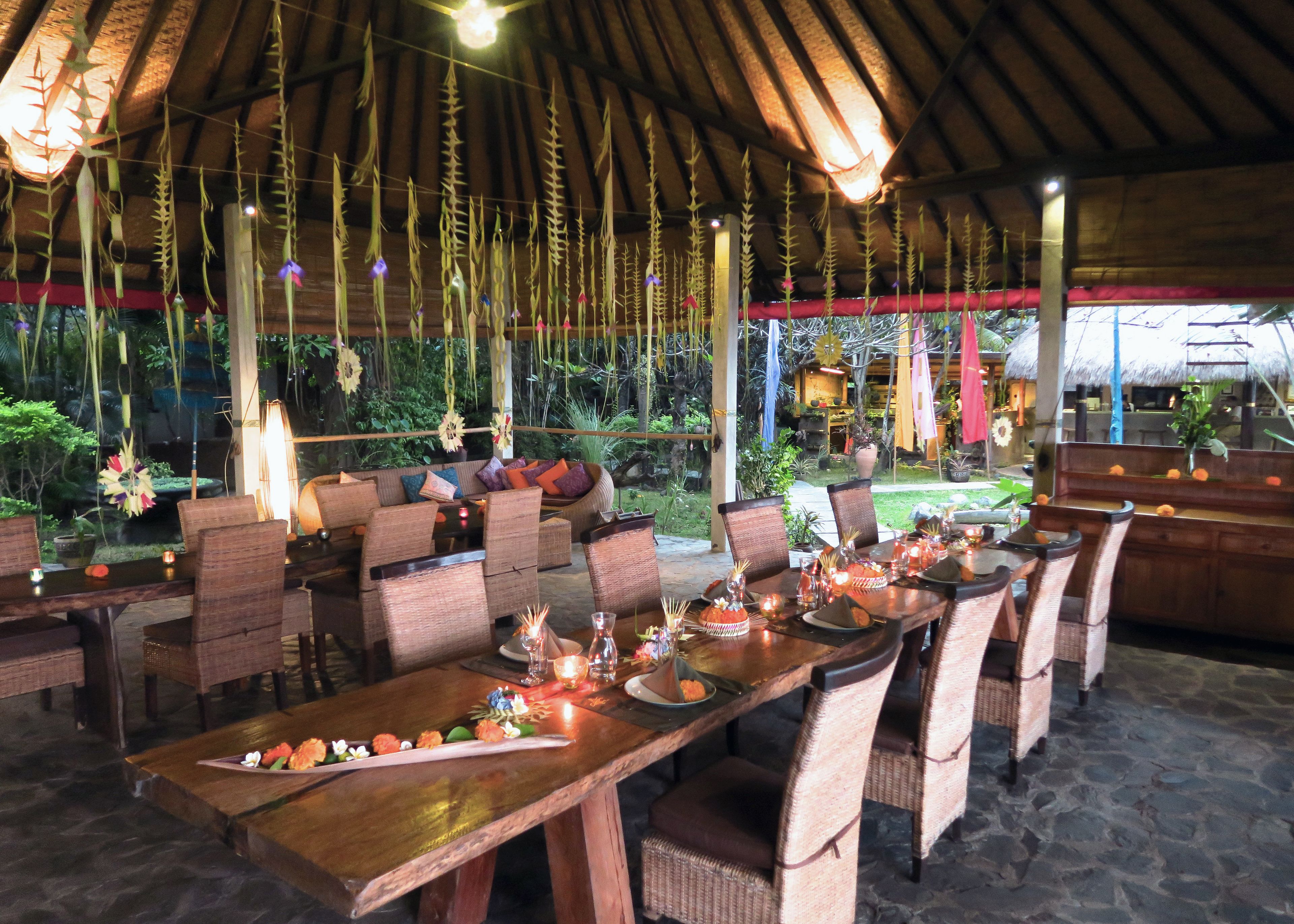 An outdoor dining area with wooden tables and chairs, surrounded by hanging plants and a thatched roof.