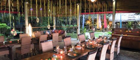 An outdoor dining area with wooden tables and chairs, surrounded by hanging plants and a thatched roof.