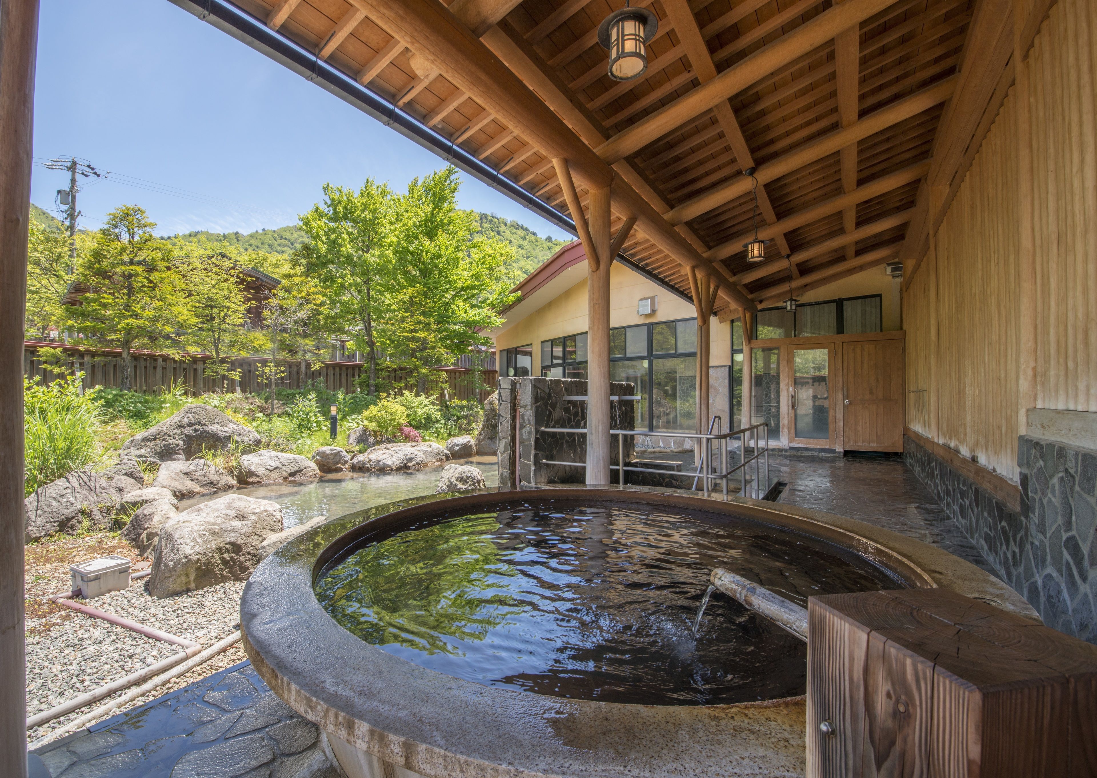 A traditional Japanese onsen with a stone bath, wooden beams, and a view of a forested hillside.