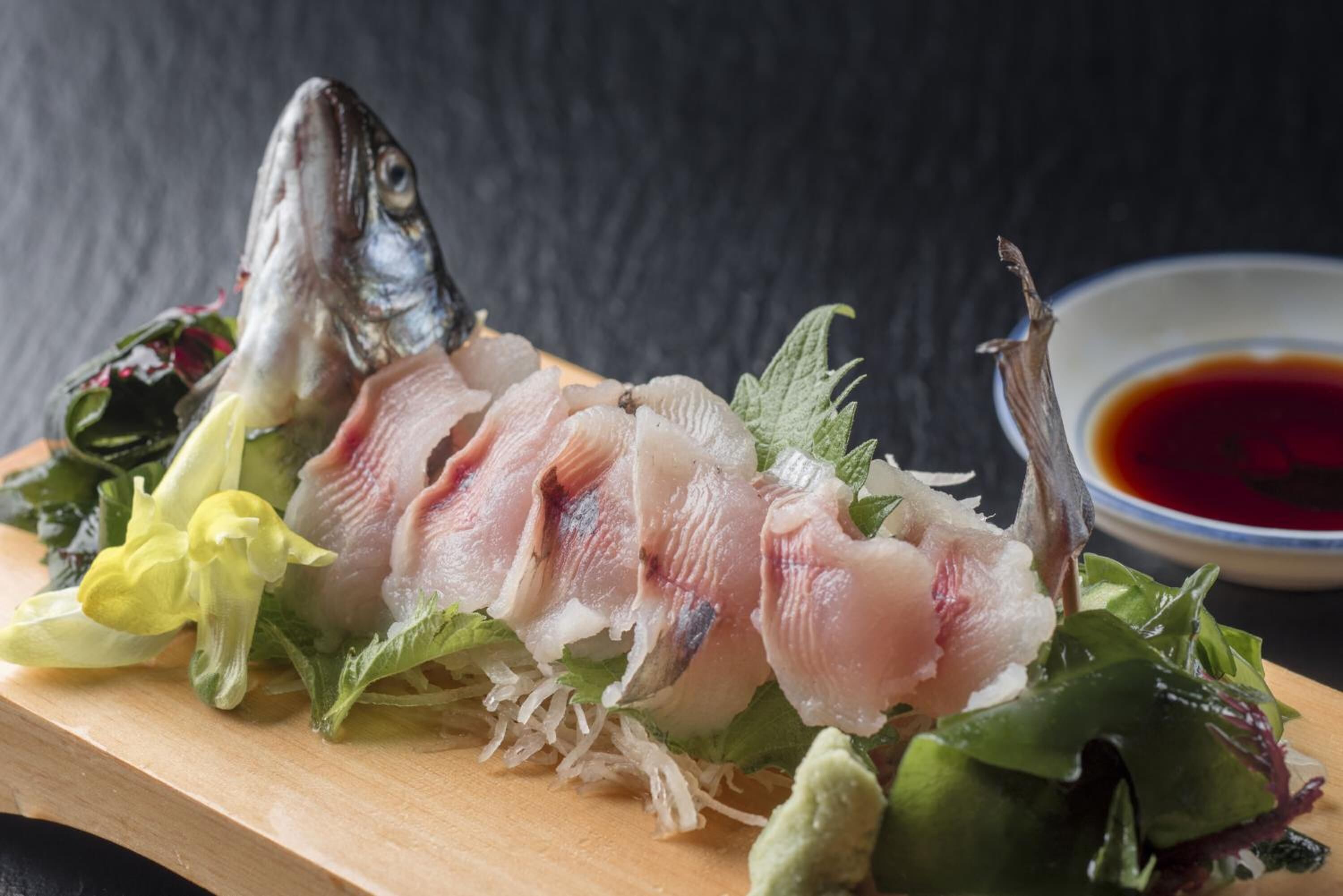Una tabla de madera con pescado en rodajas, verduras y un recipiente pequeño con salsa.