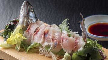 Una tabla de madera con pescado en rodajas, verduras y un recipiente pequeño con salsa.